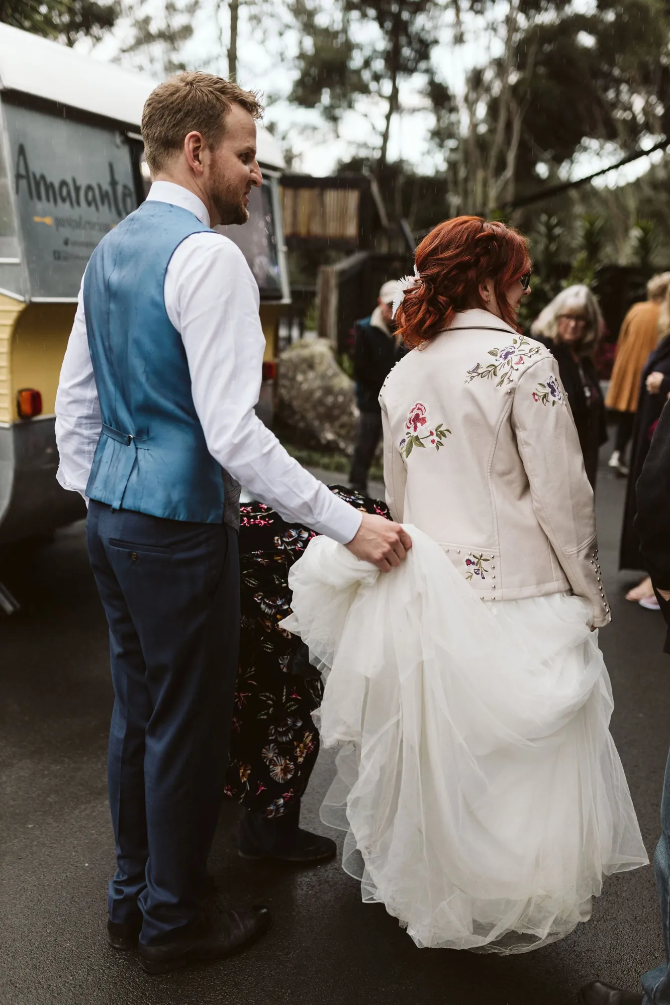 Vince holding the train of Amy's dress in the rain beside the Amaranto caravan, her cream embroidered leather jacket visible