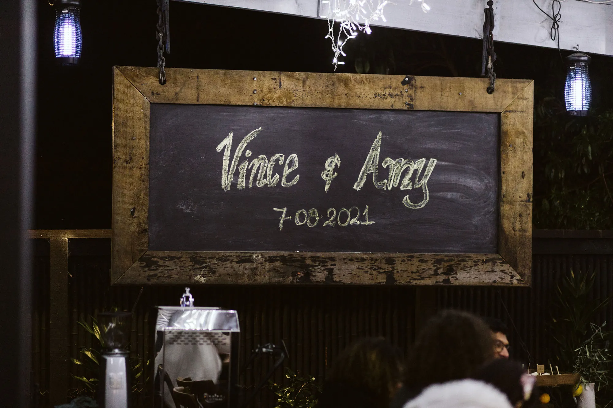 Chalkboard sign reading Vince & Amy 7.08.2021 hanging under fairy lights
