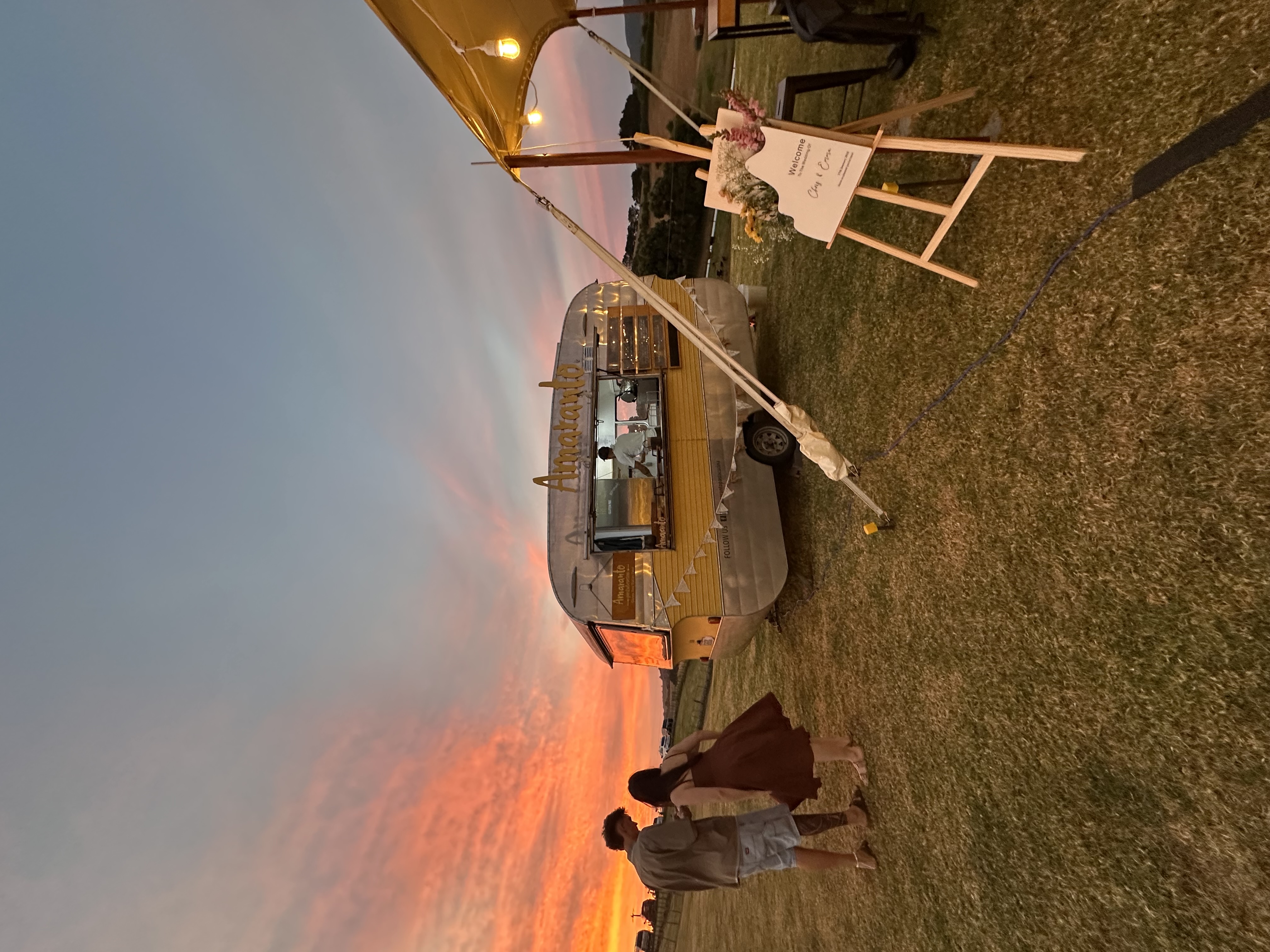 The Amaranto vintage food truck caravan at sunset during a wedding in Northland, New Zealand, with a couple in the foreground and Tommaso cooking inside