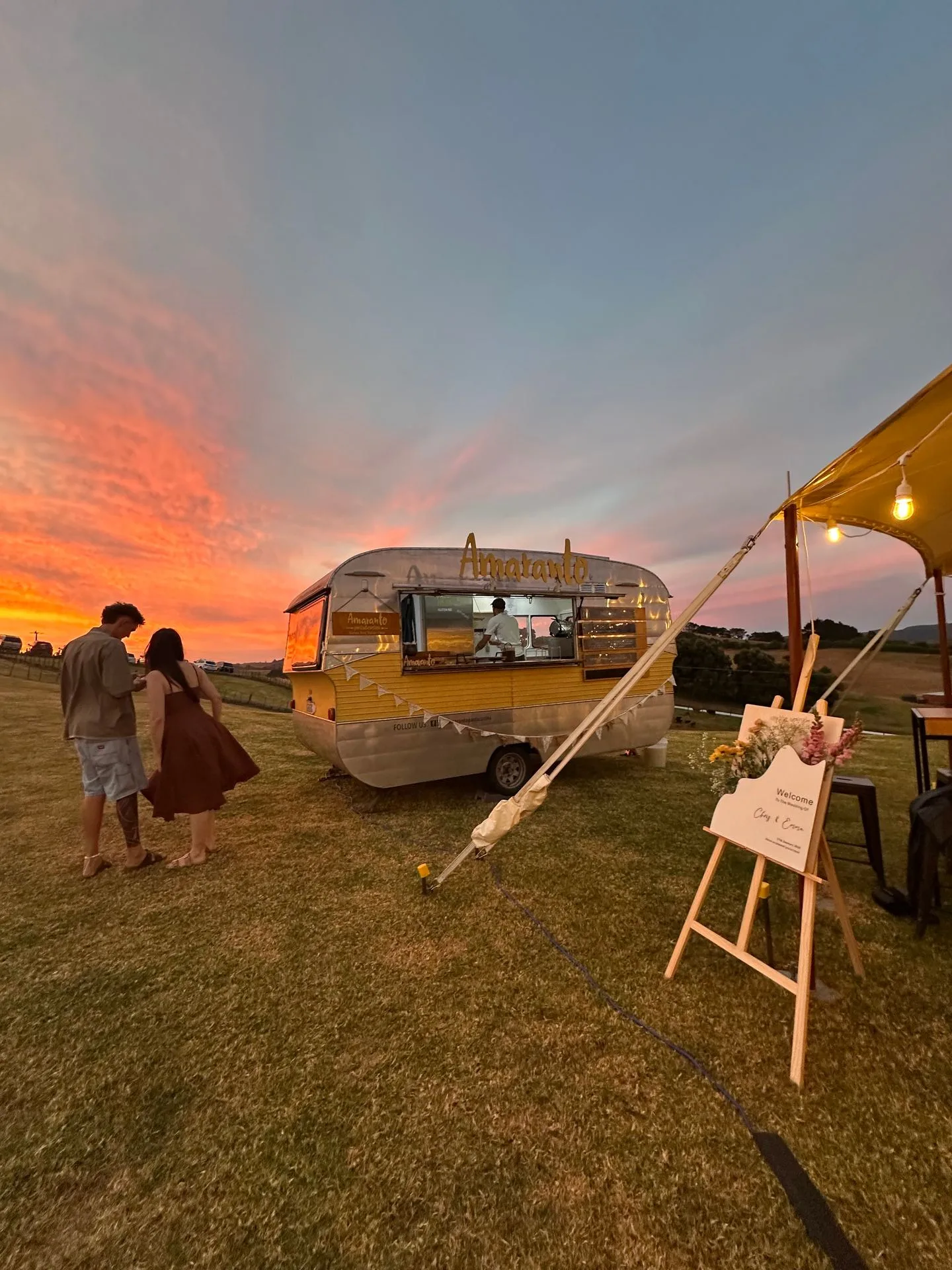 The Amaranto vintage food truck caravan at sunset during a wedding in Northland, New Zealand, with a couple in the foreground and Tommaso cooking inside