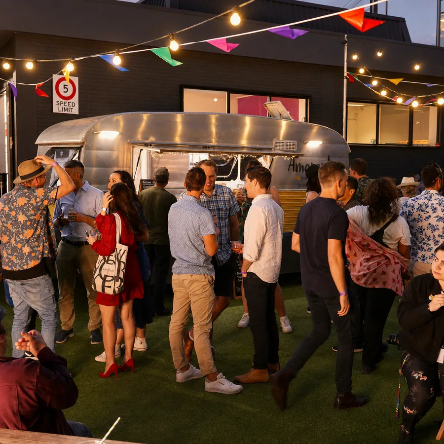 Guests mingling at an Amaranto catered evening corporate event in Auckland, with the vintage silver caravan lit up in the background, colourful bunting and fairy lights strung overhead