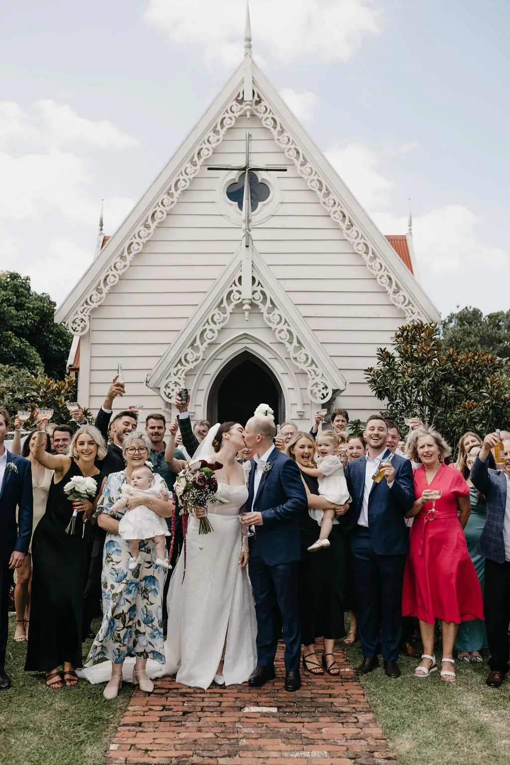 Chloe and Mitchell kissing outside the Matakana village chapel as guests cheer and raise their glasses