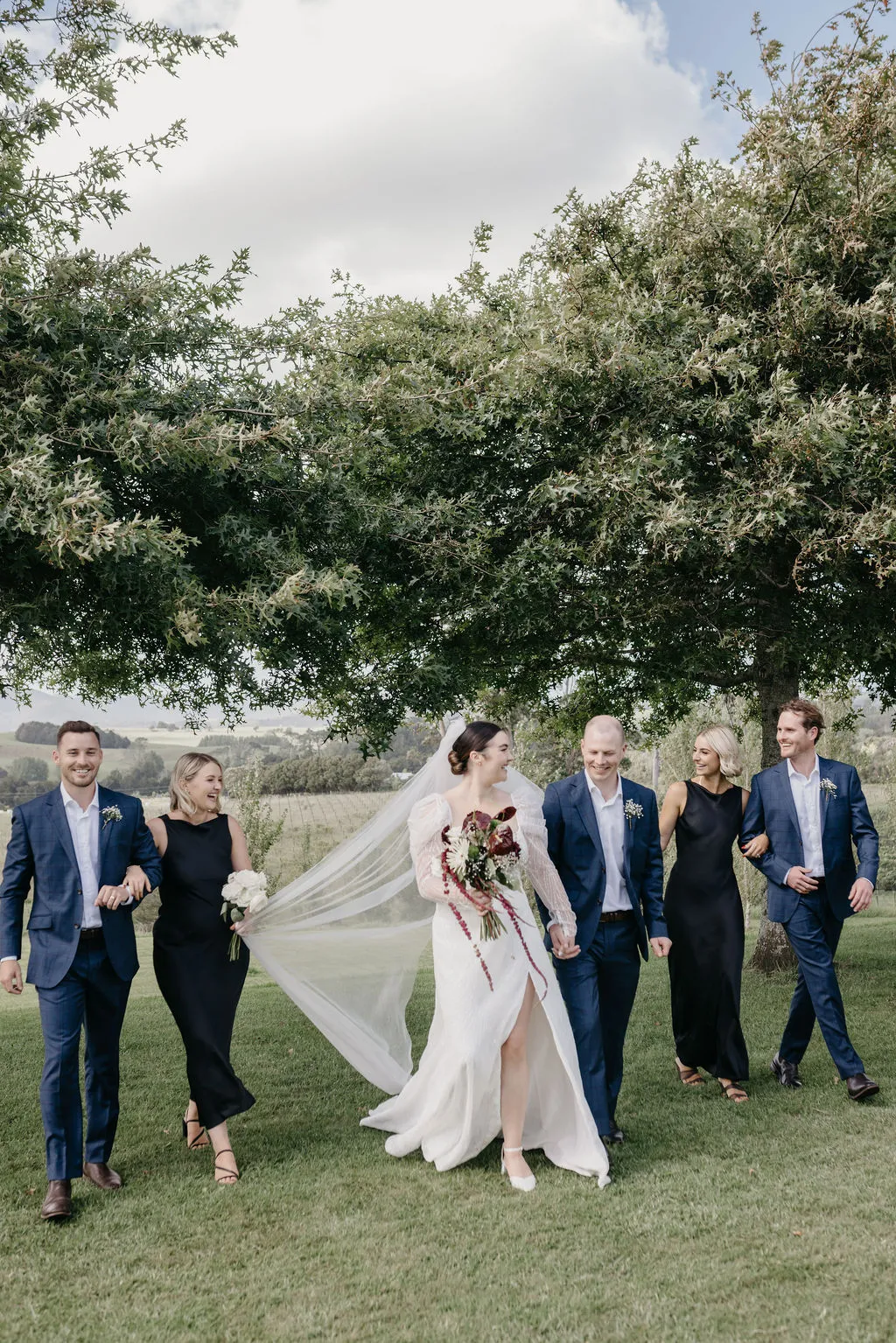 Chloe and Mitchell's wedding party walking across the Matakana fields, veil flying in the wind