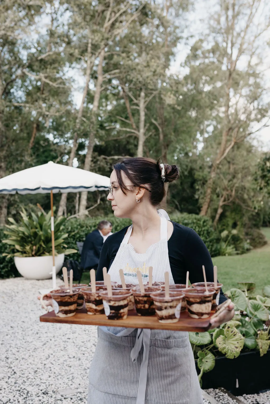 An Amaranto staff member carrying a tray of tiramisu cups through the garden at Six One Five Matakana