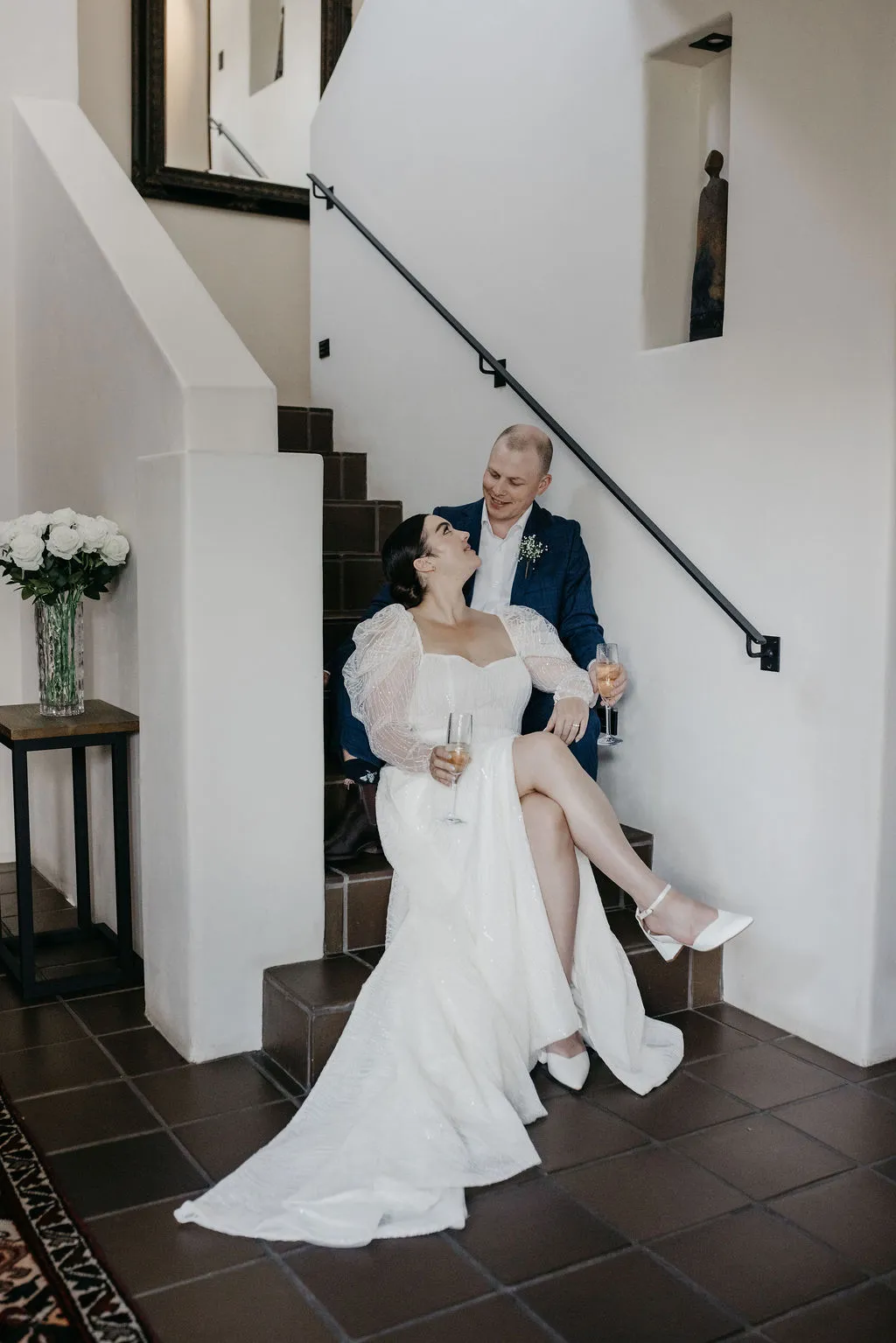 Chloe and Mitchell sharing a laugh on the stairs with glasses of champagne at Six One Five Matakana