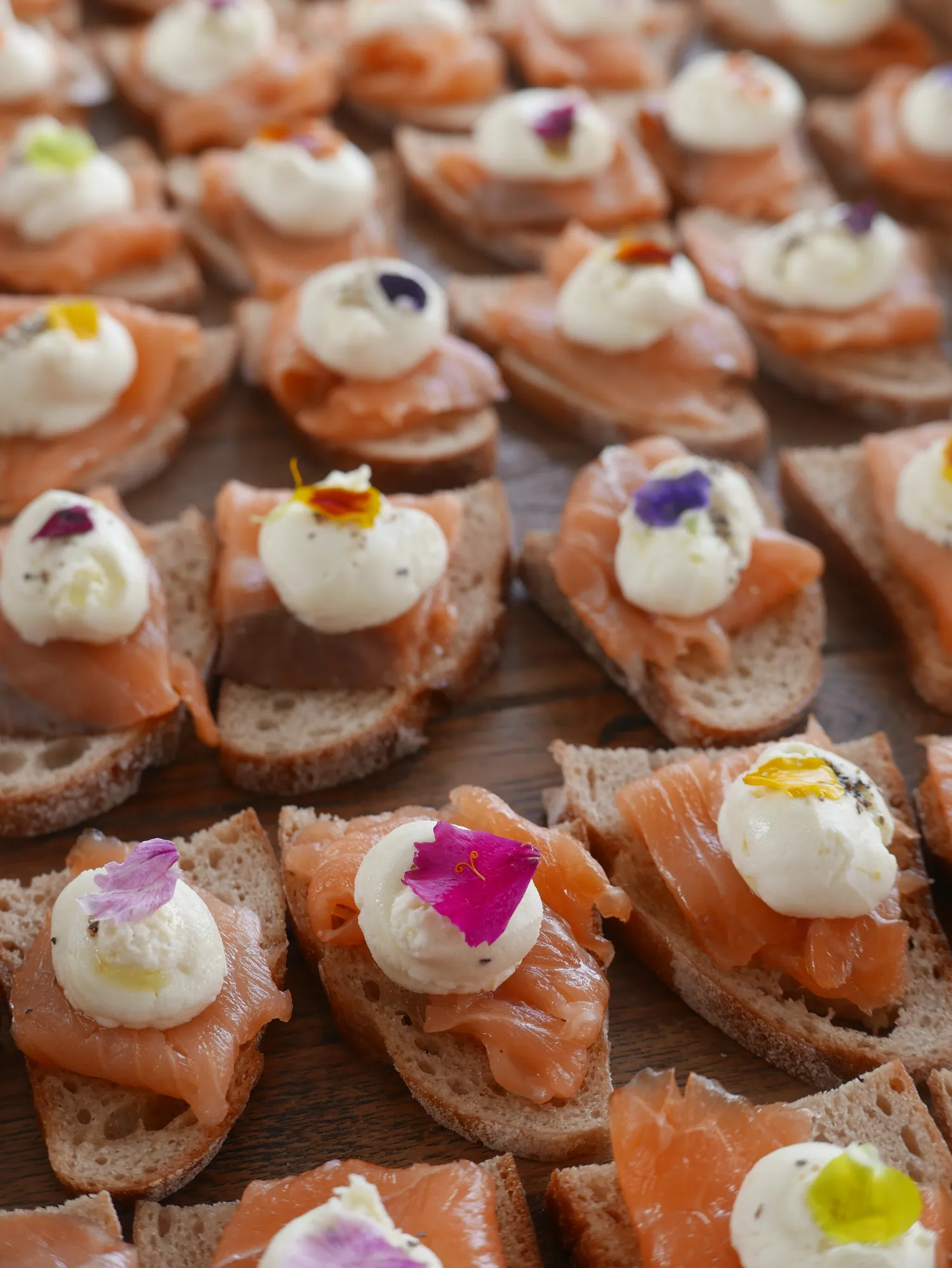 Rows of smoked salmon canapés on bread with whipped cream cheese and edible flowers, prepared by Amaranto for a wedding.