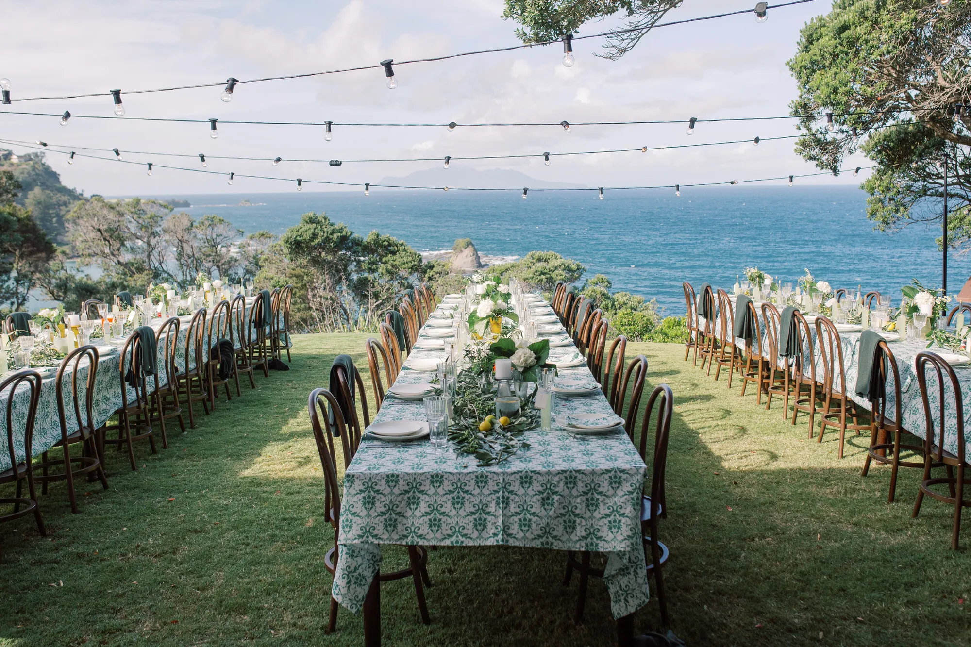 Wide al fresco tablescape with long tables, green damask cloths and ocean views stretching behind at Elle and Sam's wedding at Ti Point, Auckland