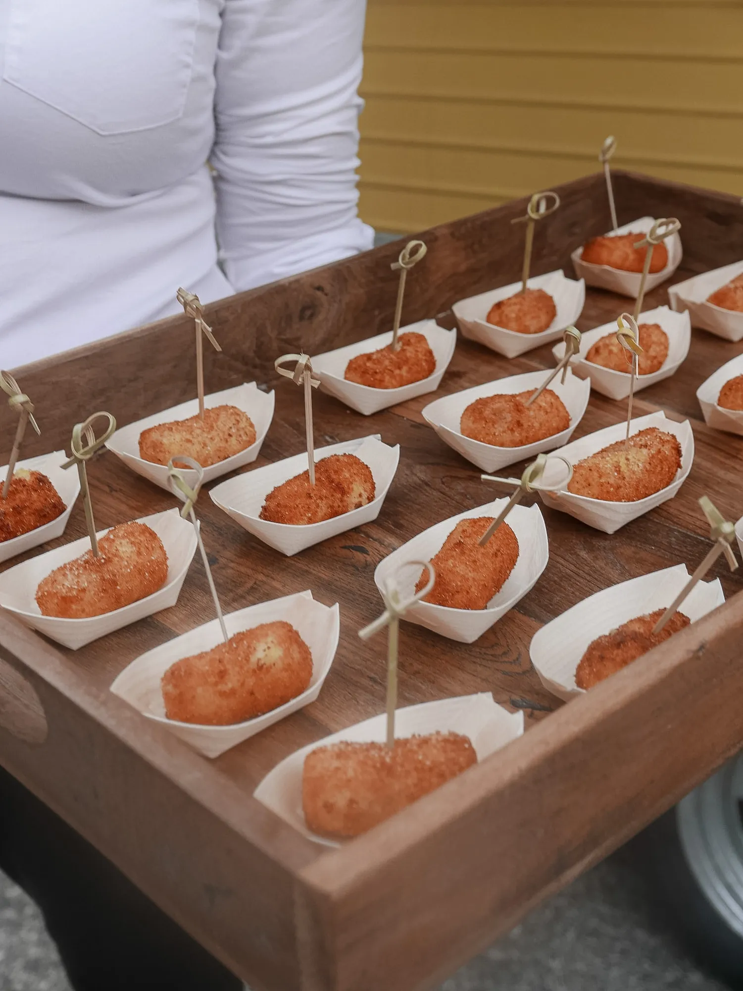 Golden potato and prosciutto crocchette in paper boat trays on a wooden tray, with the yellow Amaranto caravan visible behind