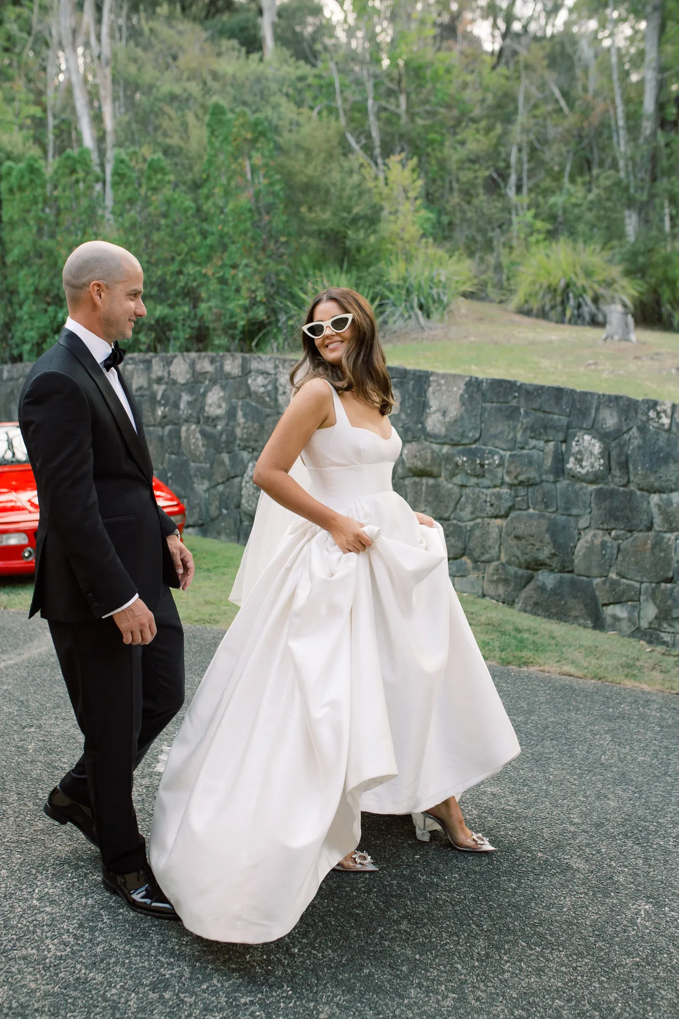 Elle in sunglasses walking with Sam on the sunny grounds at Ti Point on their wedding day