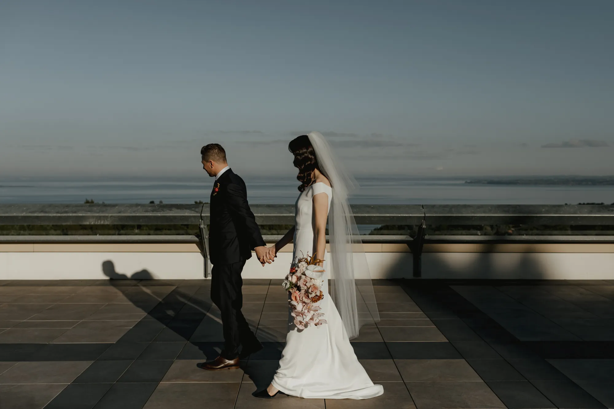 Lily and Dylan holding hands on the terrace at French Bay Yacht Club with the sea behind them