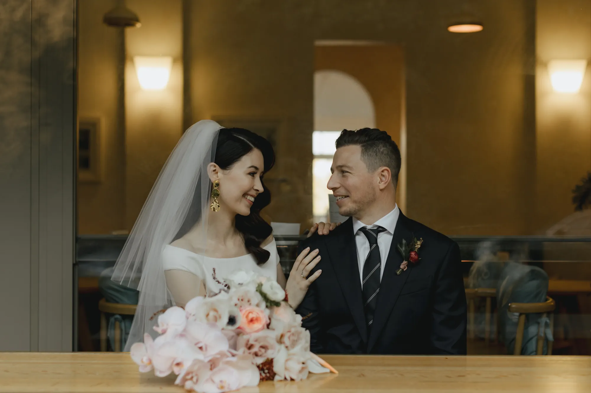 Lily and Dylan smiling at each other through a window, her bouquet resting on the bench
