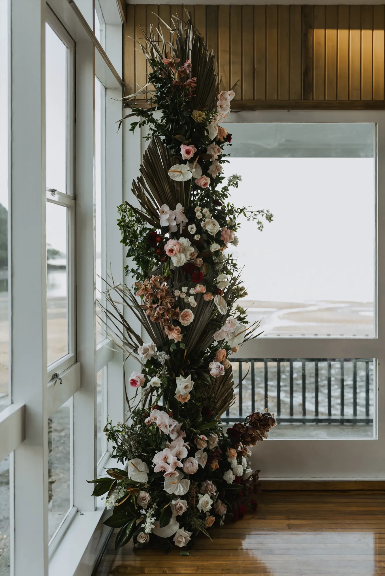 Tall floral installation of roses, orchids, anthurium and dried palm by a window overlooking the bay