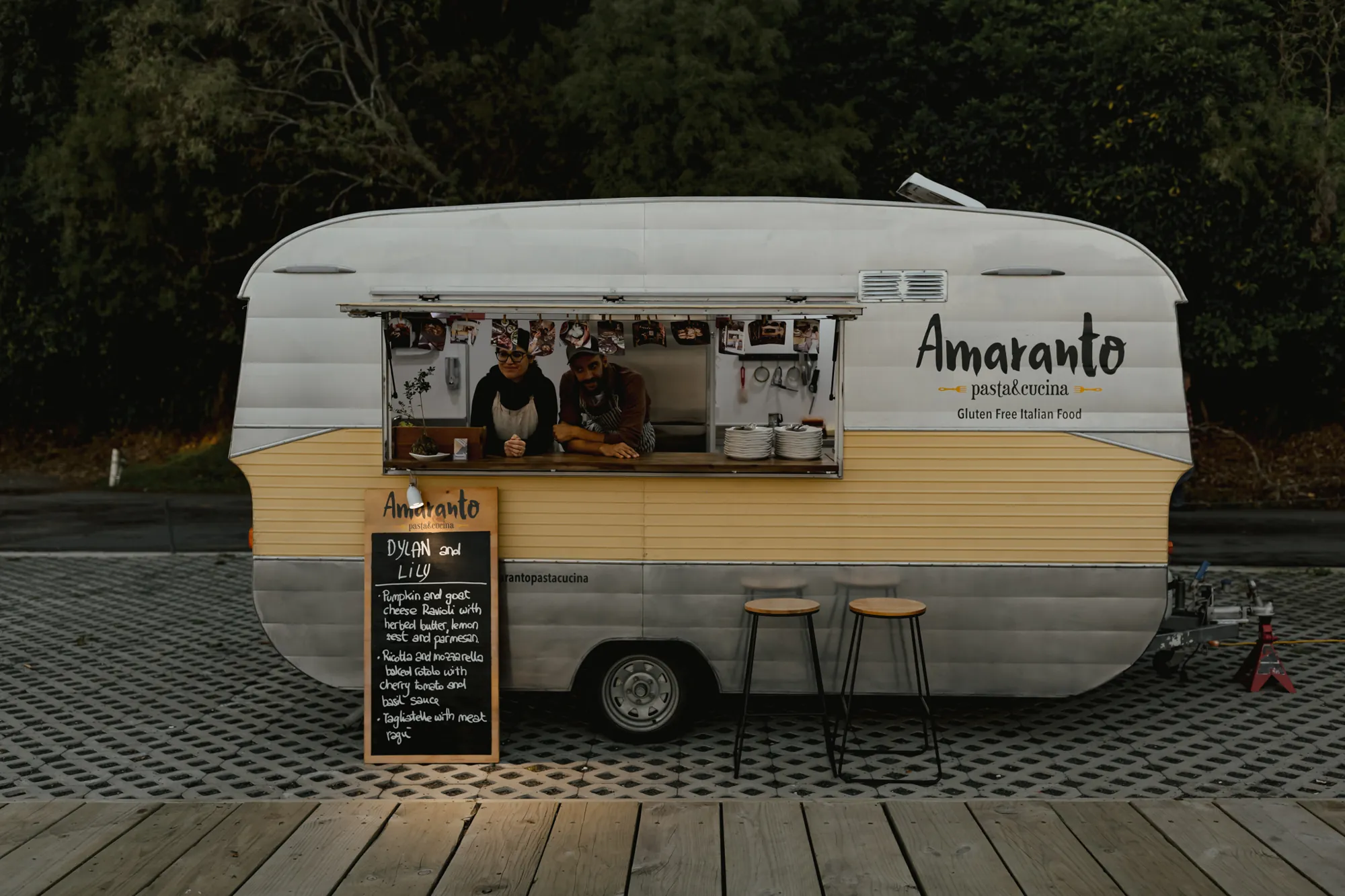 The Amaranto Pasta & Cucina caravan at dusk with Tommy and Luisa in the window and a chalkboard menu for Dylan and Lily