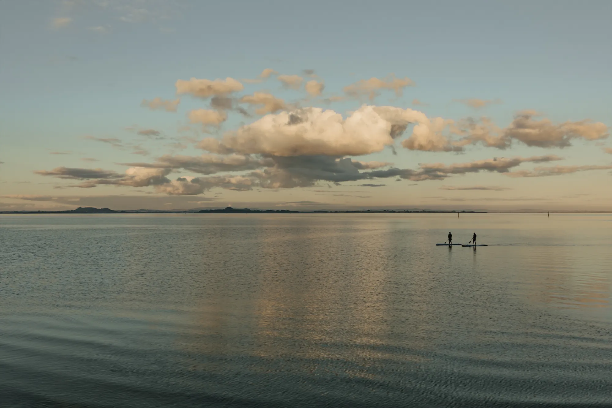 Two paddleboarders on a calm bay at dusk under a large cloud
