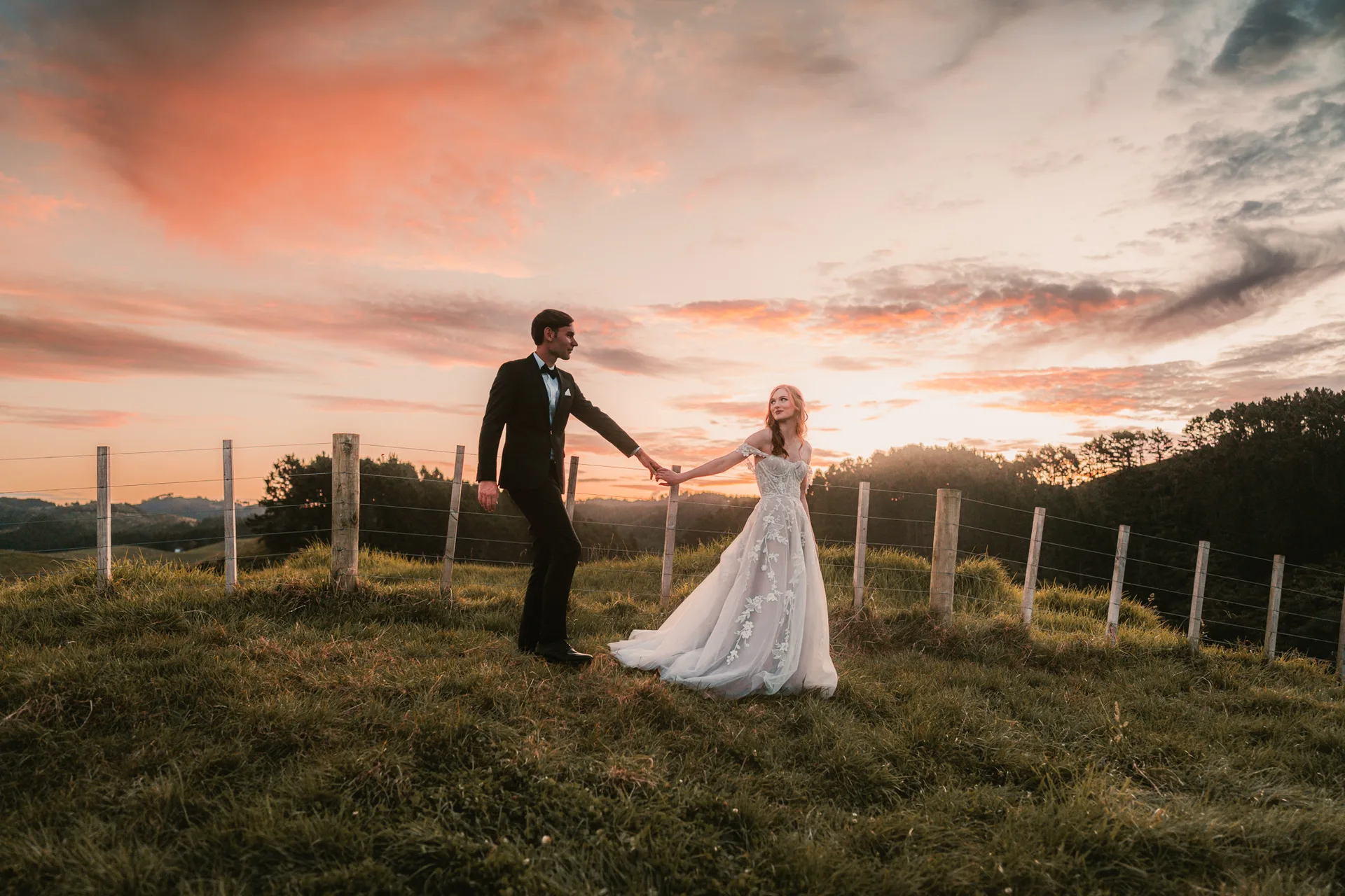 Tim and Courtney holding hands on the hilltop at Nocton Woolshed under a dramatic orange and pink sunset sky, with the rolling Northland landscape stretching to the horizon