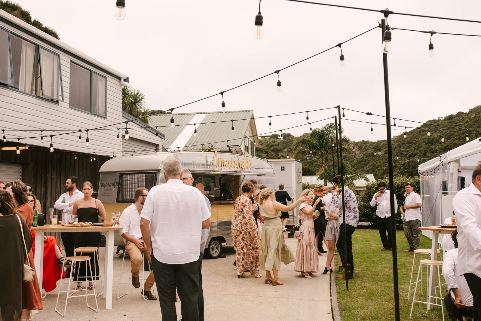 Guests mingling around the Amaranto caravan under festoon lights at Sandi and Ryan's wedding, Whangaumu Bay